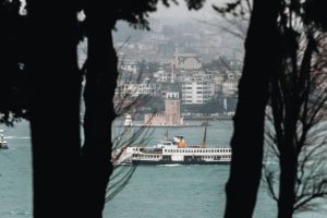 A serene view of a ferry on the Bosphorus with Istanbul skyline in the background, framed by trees.